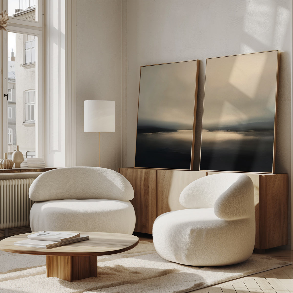 A cozy, modern living room features two white rounded armchairs, a low wooden coffee table, and a sideboard beneath the Golden Fog Horizon - Set Of 2 wall art. Sunlight pours in through tall windows, highlighting the coastal-inspired decor.