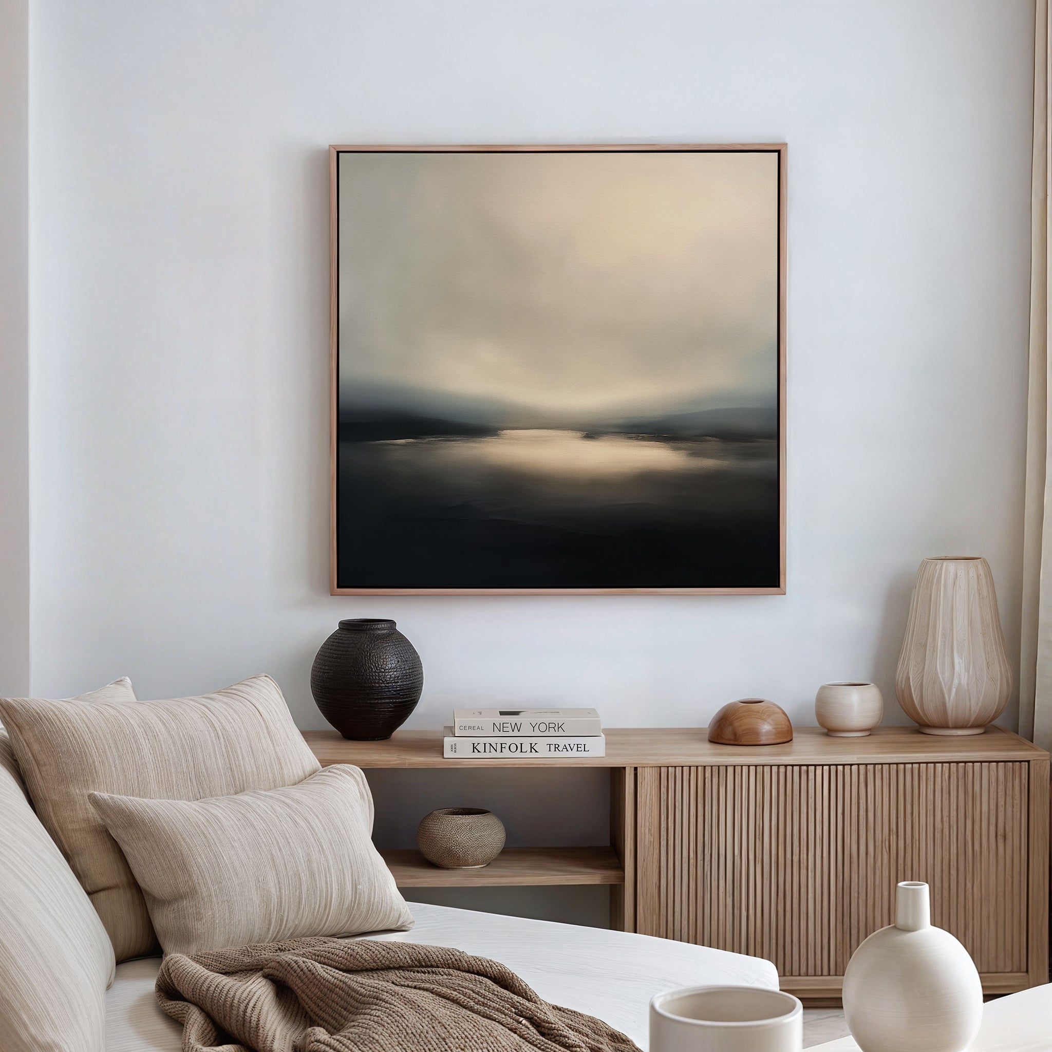 A minimalist living room featuring a light sofa, neutral pillows, a wooden sideboard topped with ceramic vases and books, and the Golden Fog Horizon Canvas Art - Square in soft, muted tones displayed above the sideboard.