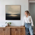 A woman in a white blouse and jeans stands by a wooden cabinet, admiring the Golden Fog Horizon Canvas Art - Square on the wall; a small potted plant and books sit neatly atop the cabinet.