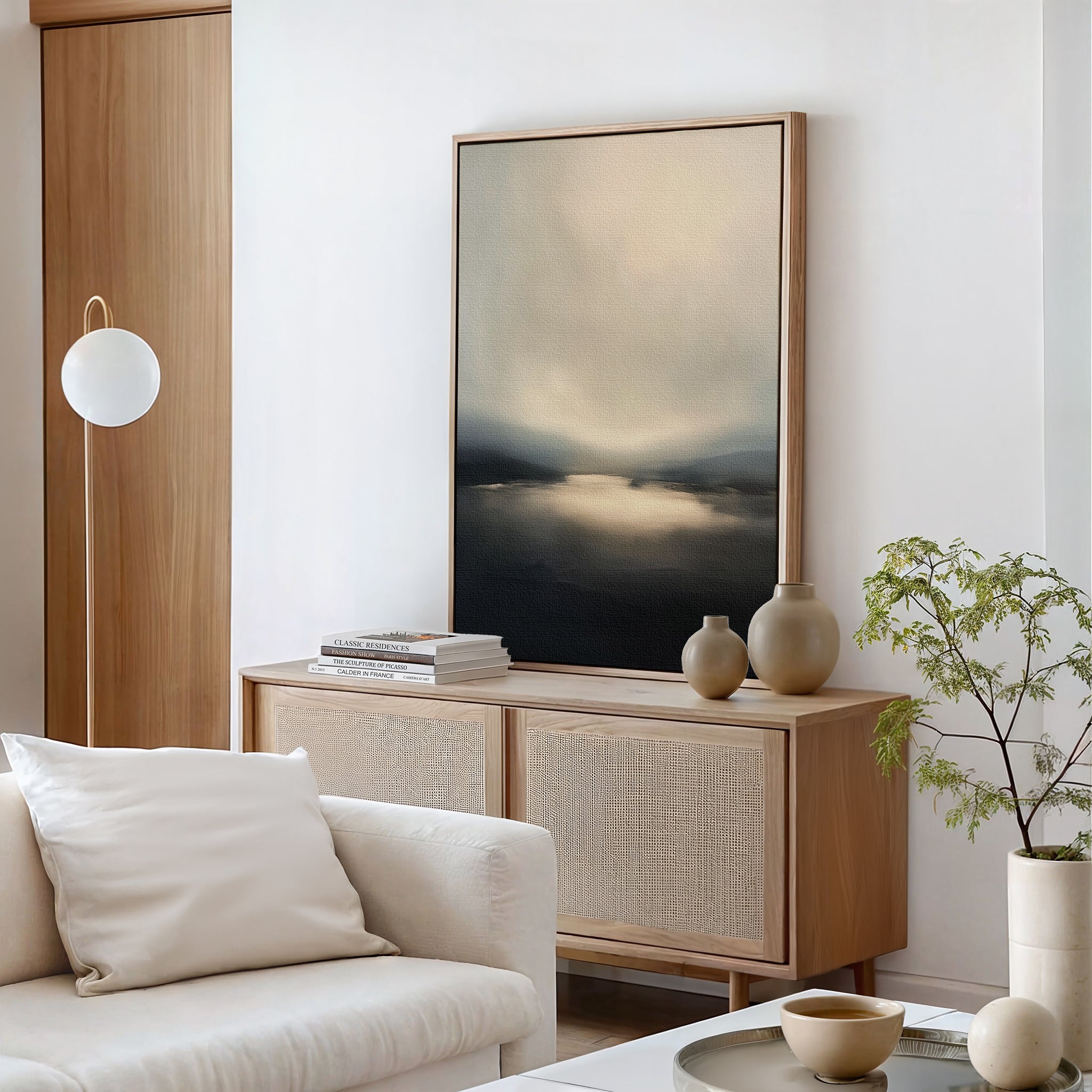 A modern living room with light wood furniture, a soft beige sofa, books, decorative vases on a sideboard, a round floor lamp, a small leafy plant, and the Golden Fog Horizon Canvas Art - Vertical creates an inviting space.