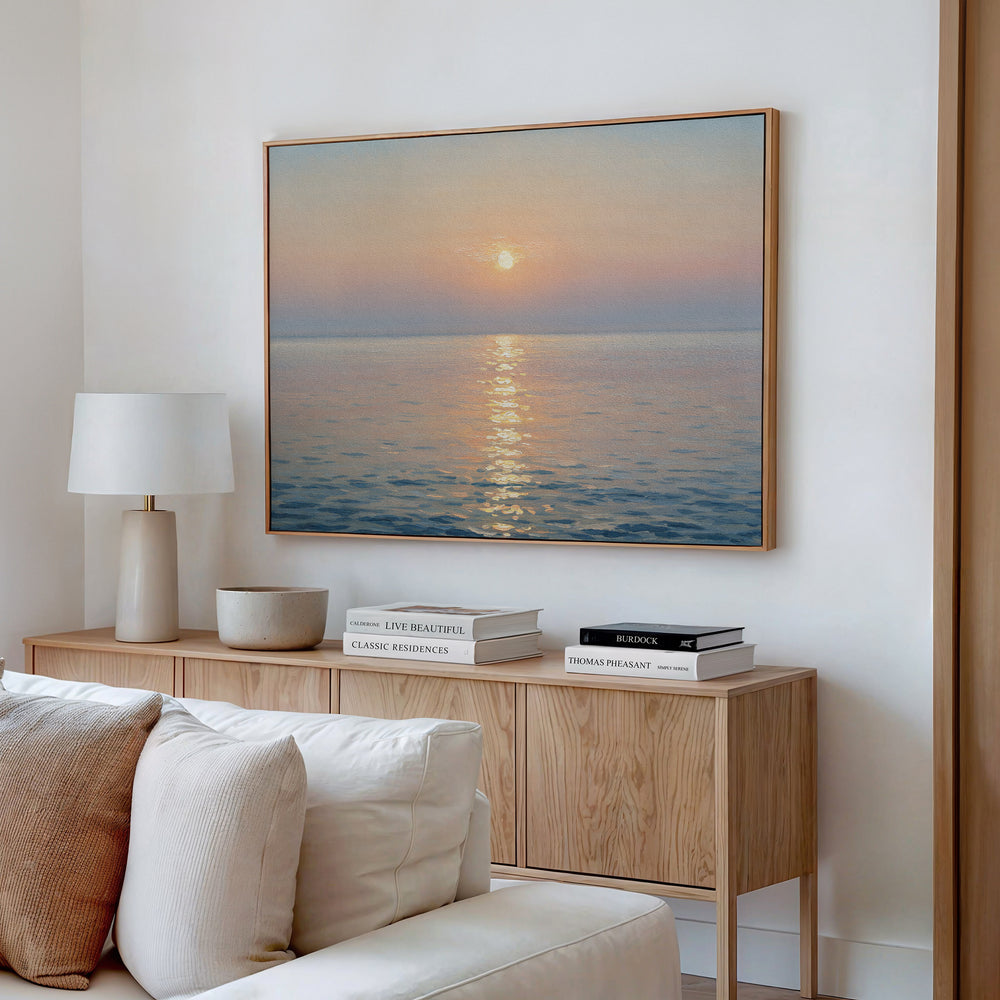 A cozy living room featuring a light wood sideboard topped with stacked books, a white table lamp, neutral decor, and serene sunset wall art in the Golden Reflection Canvas Art - Horizontal displayed above the sideboard.