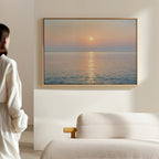 A woman in a light outfit stands in a minimalist, neutral-toned room, admiring the Golden Reflection Canvas Art - Horizontal—a serene sunset scene of calm sea and sun—hanging above a white couch, adding tranquil decor to the space.