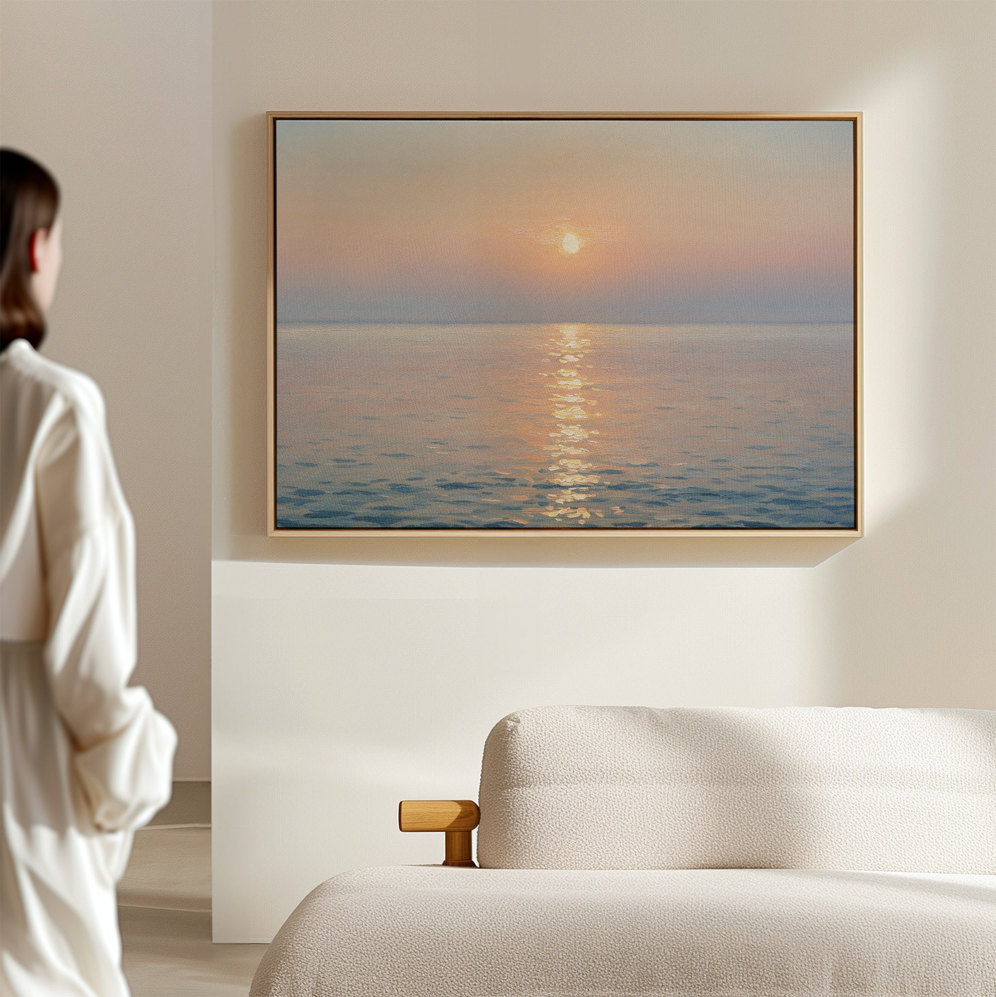 A woman in a light outfit stands in a minimalist, neutral-toned room, admiring the Golden Reflection Canvas Art - Horizontal—a serene sunset scene of calm sea and sun—hanging above a white couch, adding tranquil decor to the space.
