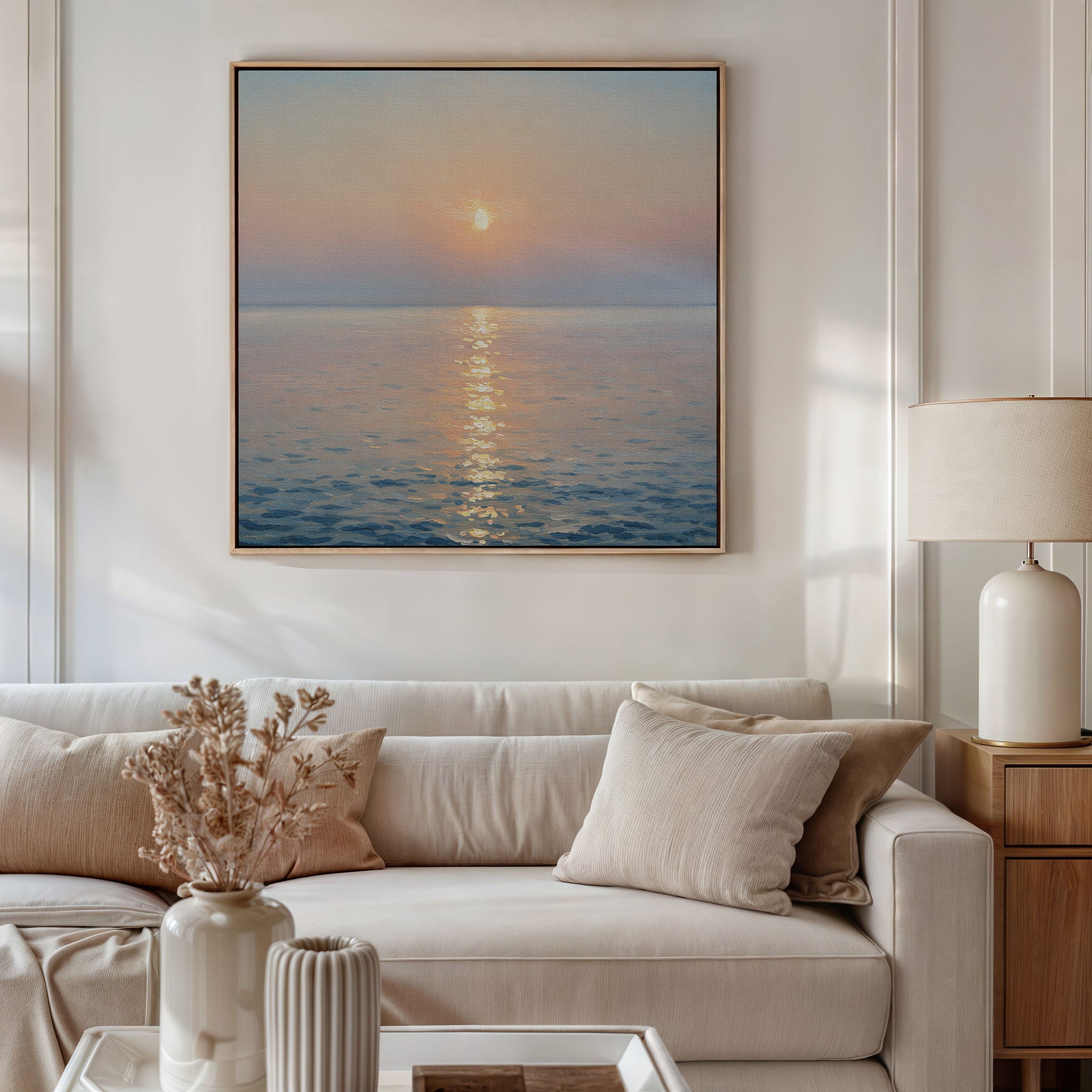 A modern living room features a beige sofa with light cushions, a side table and lamp, dried flowers in a vase, and the Golden Reflection Canvas Art - Square depicting a serene ocean sunset hanging on the wall.