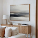 A modern living room features a light wooden sideboard with a white table lamp, stacked books, and a ceramic bowl. The Grey Ripples Canvas Art - Horizontal hangs above on the white wall, with a beige sofa in the foreground.