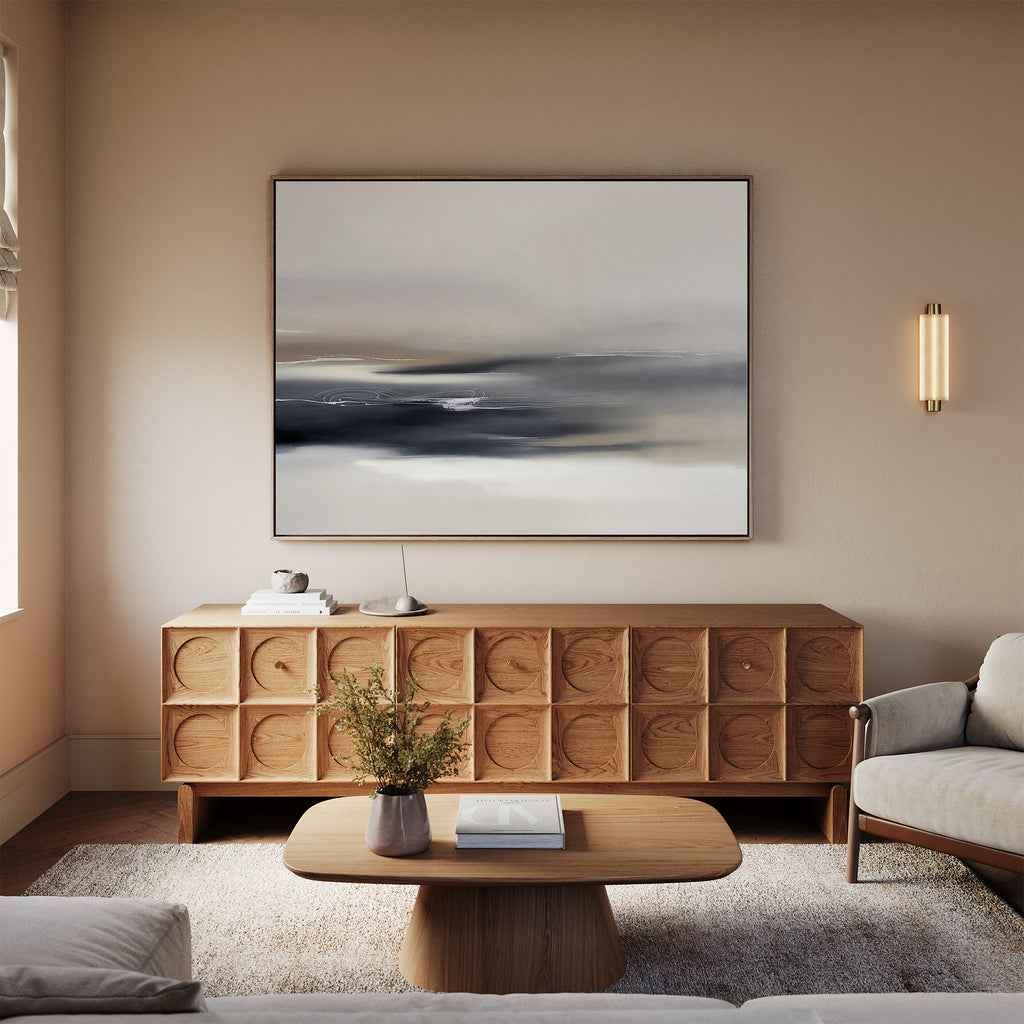 A modern living room featuring a wooden sideboard, beige sofa, wooden coffee table with a book and small vase, neutral walls, sconce light fixture, and the Grey Ripples Canvas Art - Horizontal adds a stylish touch perfect for modern design fans.