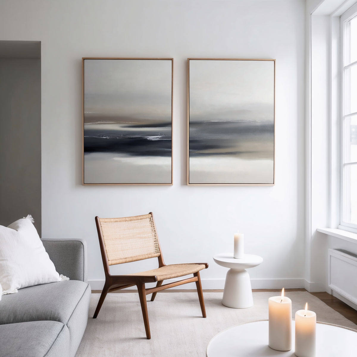 A modern, minimal living room featuring a wooden chair, small white side table with a lit candle, the Grey Ripples - Set Of 2 abstract artwork on the wall, a gray sofa with a white pillow, and more candles on a round white coffee table.