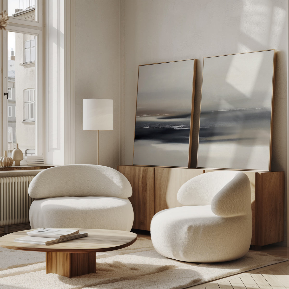 A modern living room featuring the Grey Ripples - Set Of 2 white rounded armchairs, a low wooden table, sideboard, and minimalist prints and abstract paintings, all bathed in natural light from large windows.