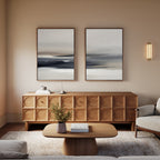 A cozy living room features a wooden sideboard with a geometric pattern, Grey Ripples - Set Of 2 artworks above it, a soft beige armchair, a wooden coffee table with a book and vase, and warm neutral walls softly lit.