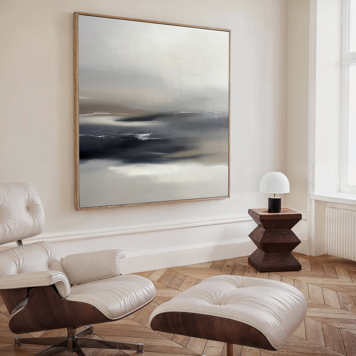 A modern, minimalist room featuring the Grey Ripples Canvas Art - Square, a white leather lounge chair and ottoman, a geometric wooden side table, and a small white lamp by the tall window—ideal for modern living spaces.