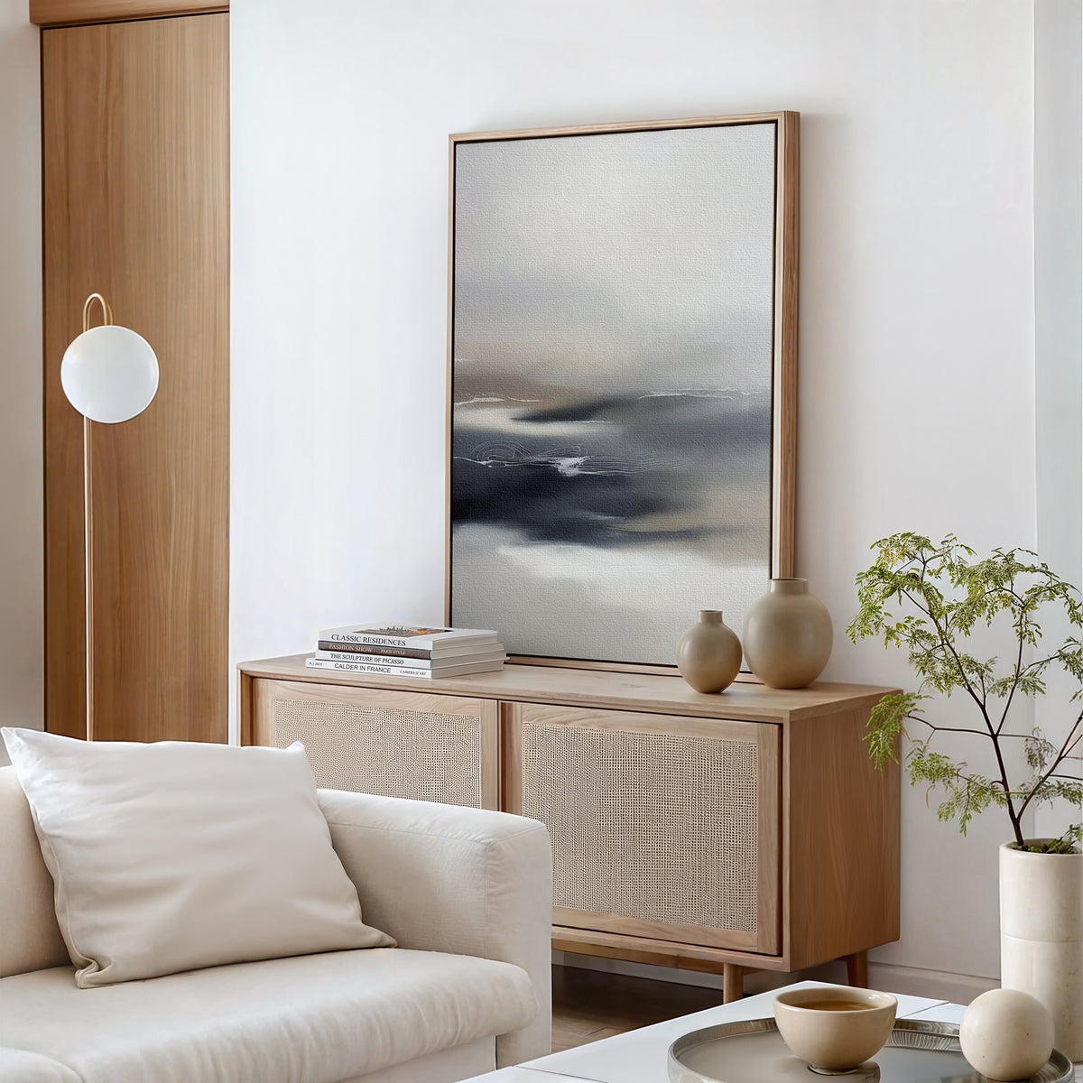 A modern living room featuring a light wood sideboard, the Grey Ripples Canvas Art - Vertical, decorative vases, stacked books, a potted plant, a white sofa with a cushion, and a round floor lamp against a white wall.