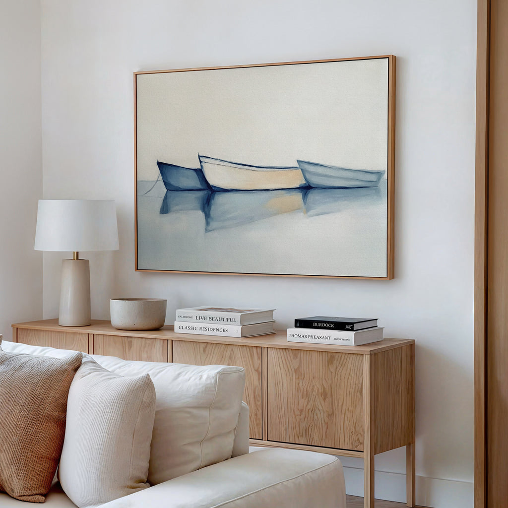 A minimalist living room features a light wood sideboard with books and a lamp, a beige sofa, and the Harbor Rest Canvas Art - Horizontal showcasing three blue and white boats on calm water for soothing decor.