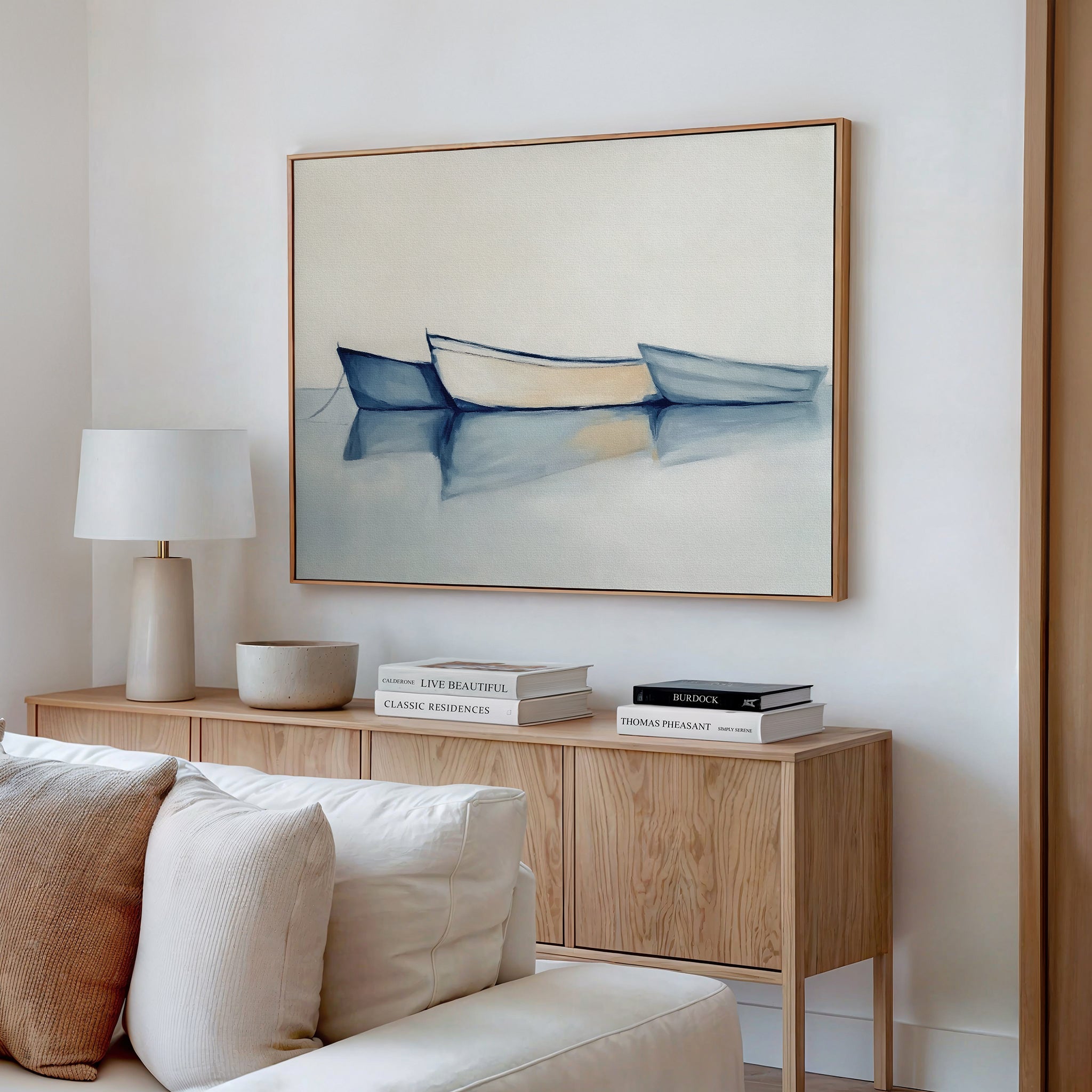A minimalist living room features a light wood sideboard with books and a lamp, a beige sofa, and the Harbor Rest Canvas Art - Horizontal showcasing three blue and white boats on calm water for soothing decor.