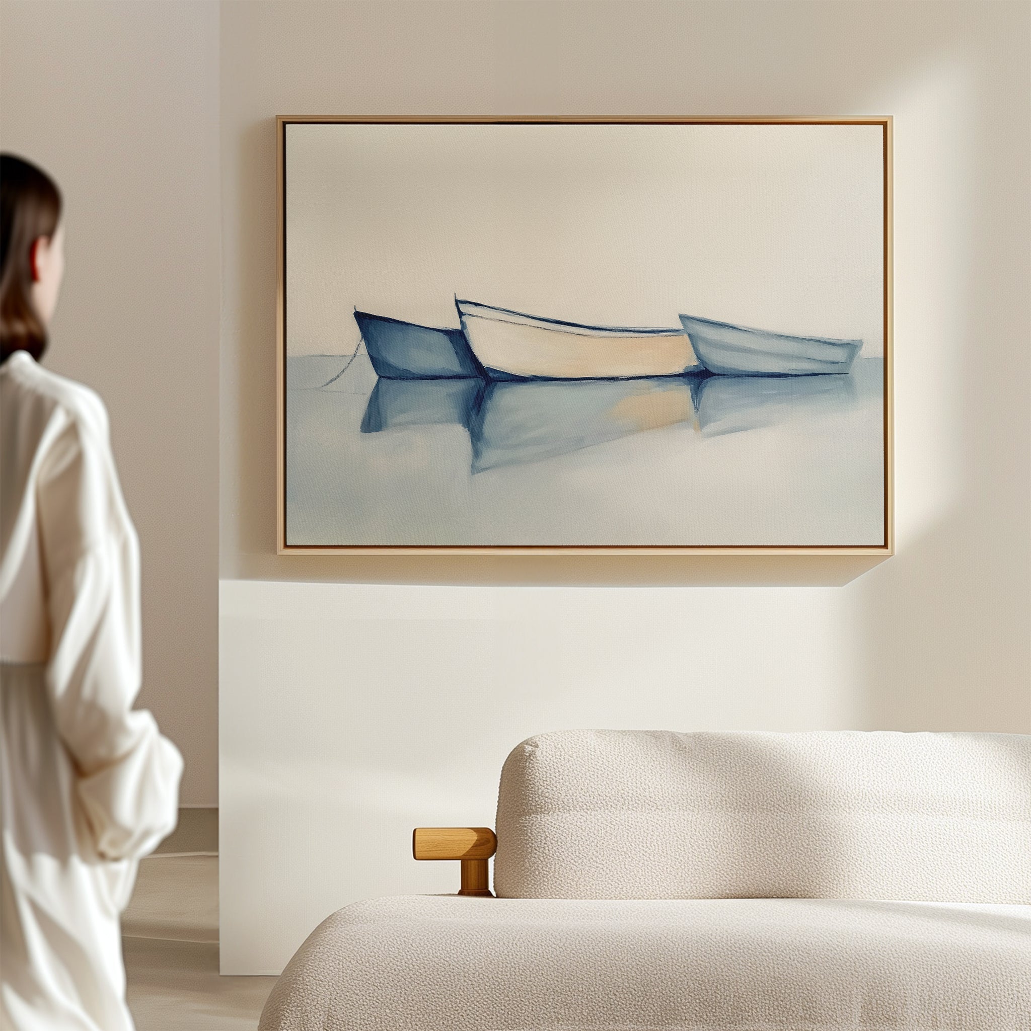 In a minimal, sunlit room with a beige sofa, a person admires Harbor Rest Canvas Art—horizontal wall decor featuring three blue and white rowboats on tranquil water—ideal as calming coastal or maritime-inspired decor.