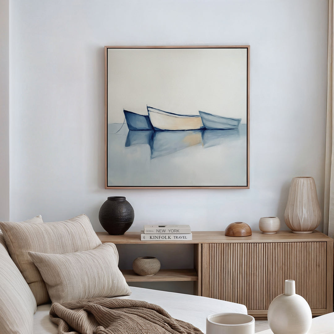 A modern living room with a beige sofa, neutral cushions, and a wooden sideboard topped with vases and books. Above, the Harbor Rest Canvas Art - Square—a painting of three boats on calm water—adds a calming coastal touch.