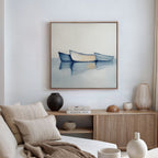A modern living room with a beige sofa, neutral cushions, and a wooden sideboard topped with vases and books. Above, the Harbor Rest Canvas Art - Square—a painting of three boats on calm water—adds a calming coastal touch.