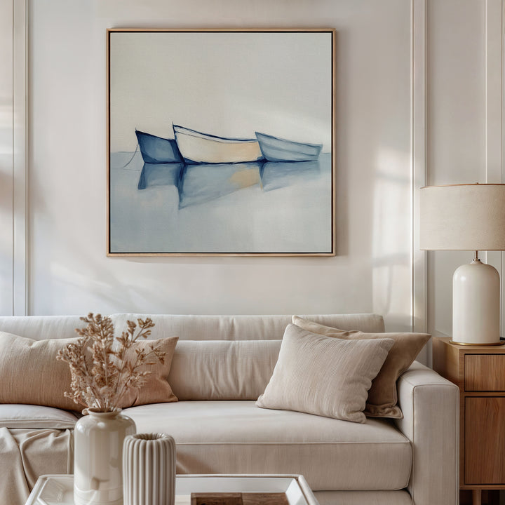 A neutral-toned living room with a white sofa, beige cushions, dried flowers, and a light wood side table topped by a ceramic lamp. Above the sofa hangs Harbor Rest Canvas Art - Square, featuring three boats on serene water.