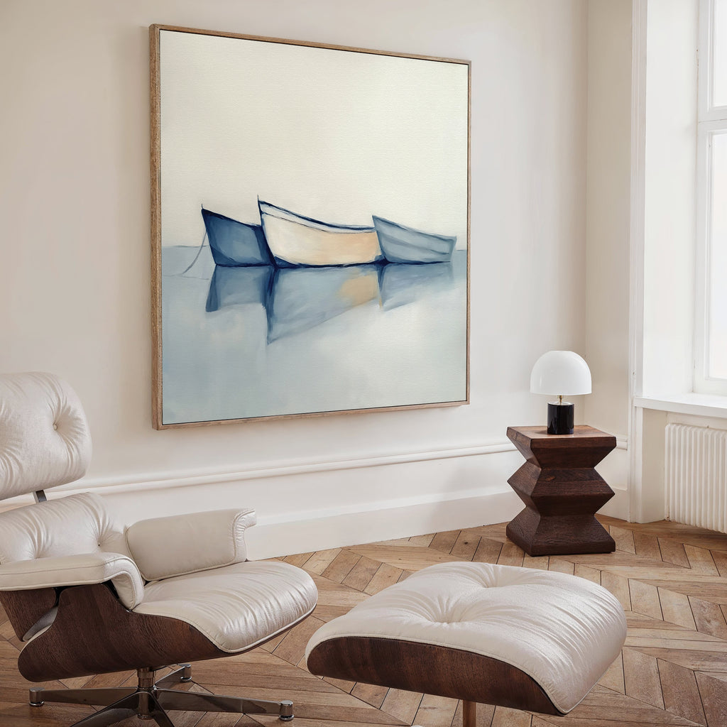 A modern, minimal living room with a white lounge chair and ottoman, wooden side table with a white lamp, and calming decor featuring the Harbor Rest Canvas Art - Square, a coastal painting of three boats on calm water hanging on the wall.
