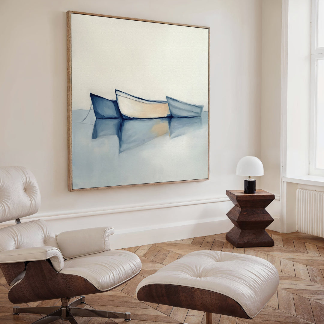A modern, minimal living room with a white lounge chair and ottoman, wooden side table with a white lamp, and calming decor featuring the Harbor Rest Canvas Art - Square, a coastal painting of three boats on calm water hanging on the wall.