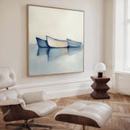 A modern, minimal living room with a white lounge chair and ottoman, wooden side table with a white lamp, and calming decor featuring the Harbor Rest Canvas Art - Square, a coastal painting of three boats on calm water hanging on the wall.