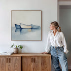 A woman in a white blouse and jeans leans on a wooden cabinet, gazing at the Harbor Rest Canvas Art - Square, featuring three boats on calm water. A potted plant and books enhance the serene decor on the cabinet below.