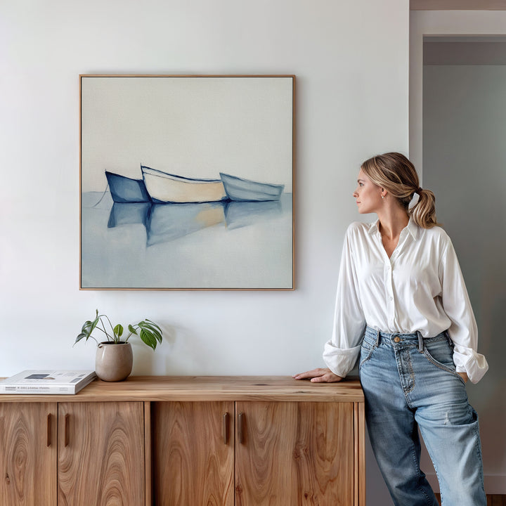 A woman in a white blouse and jeans leans on a wooden cabinet, gazing at the Harbor Rest Canvas Art - Square, featuring three boats on calm water. A potted plant and books enhance the serene decor on the cabinet below.