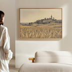 A woman stands in a softly lit room, admiring the Harvest Chapel Canvas Art - Horizontal, featuring golden fields and a distant village with a church steeple, displayed above a light-colored sofa.