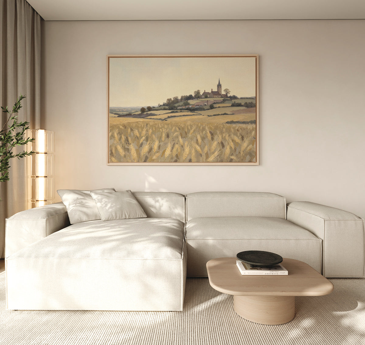 A modern living room with a light beige sectional, low wooden coffee table, and rustic Harvest Chapel Canvas Art - Horizontal above—a landscape of a village and wheat field. A tall plant and sunlight warm this cozy, inviting space.