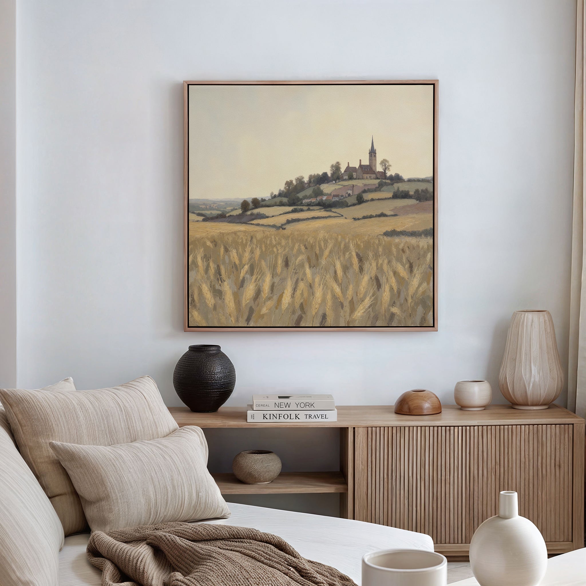 A cozy, neutral-toned living room with a beige sofa, pillows, and wood sideboard exudes rustic charm. Above the sideboard hangs the Harvest Chapel Canvas Art - Square depicting wheat fields and a village, complemented by ceramic vases.