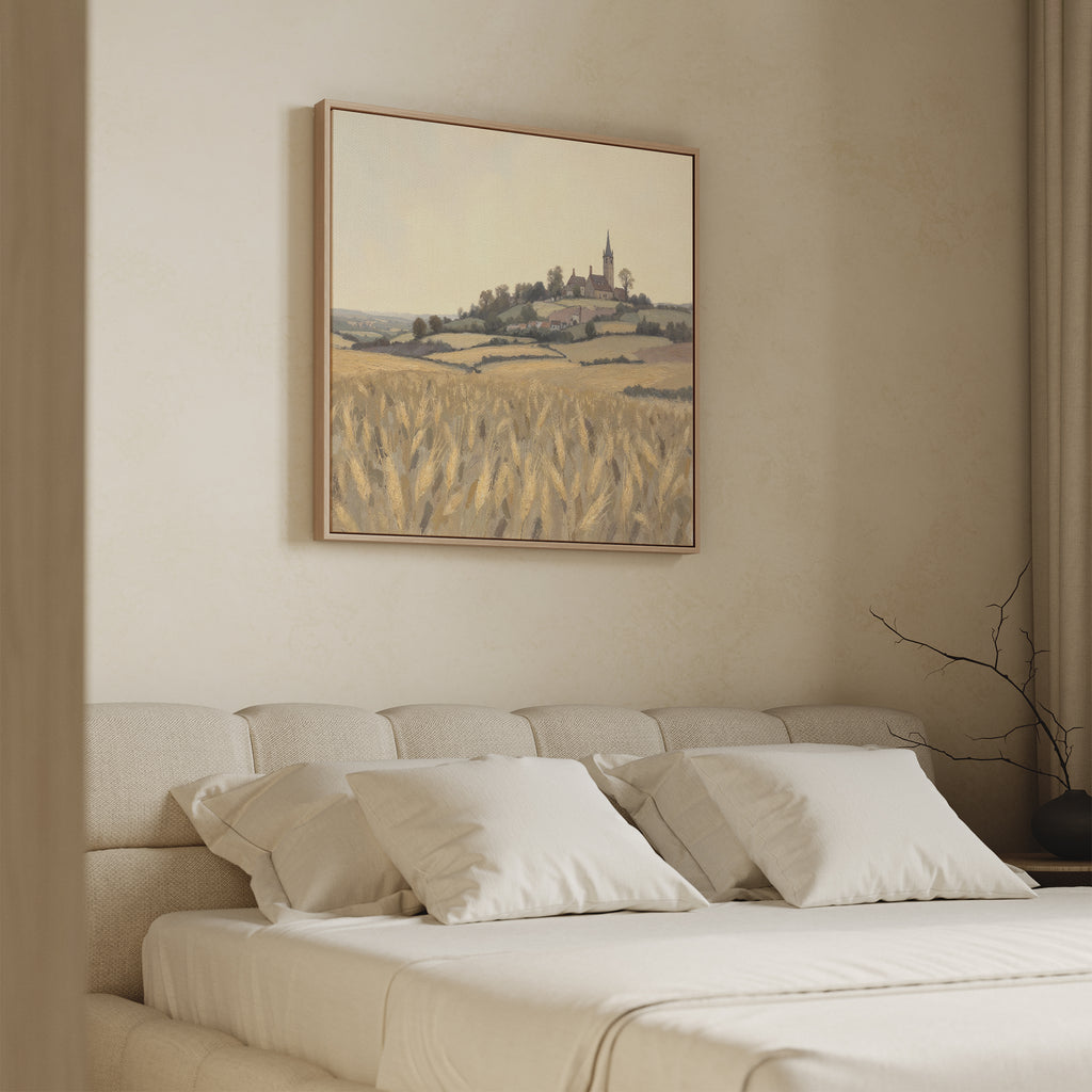 A cozy bedroom with a beige upholstered bed and white pillows, highlighted by the Harvest Chapel Canvas Art - Square above the bed, which depicts a hillside village and wheat fields for a rustic touch.