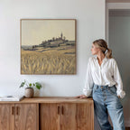 A woman in a white blouse and jeans leans against a wooden sideboard, admiring the Harvest Chapel Canvas Art - Square on the wall. A plant and books enhance the rustic charm of the bright, modern room.