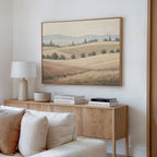 A cozy living room corner features a light wood sideboard, white lamp, decorative bowl, stacked books, and the Harvest Fields Canvas Art - Horizontal depicting fields and distant trees above. A beige sofa sits in the foreground.
