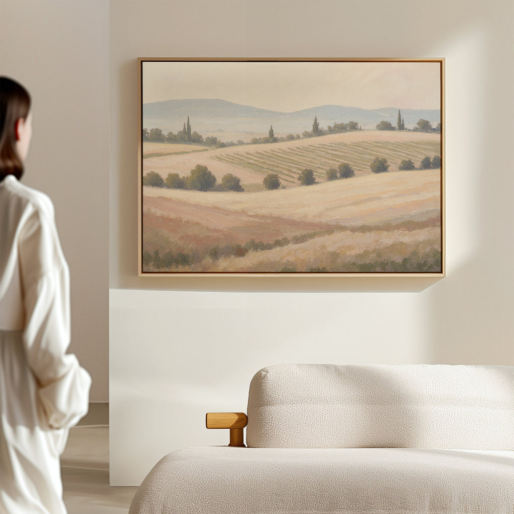 A person in a white robe stands in a minimal, light-filled room, admiring the Harvest Fields Canvas Art - Horizontal depicting rolling fields and distant hills above a beige upholstered sofa.