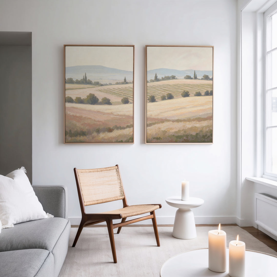 A modern minimalist living room featuring a woven chair, white side table, candles, gray sofa, and Harvest Fields - Set of 2 canvas paintings on a white wall, with natural light streaming through large windows.