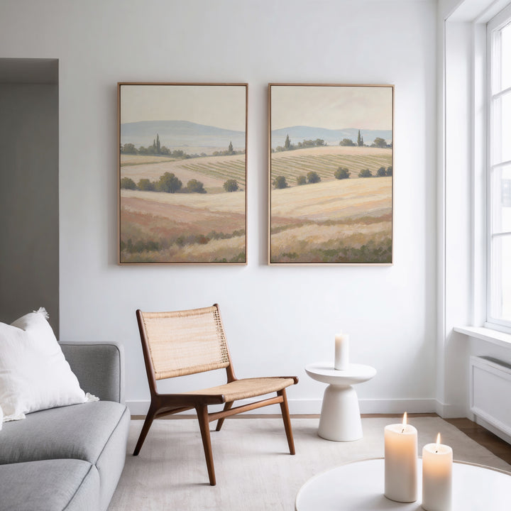 A modern minimalist living room featuring a woven chair, white side table, candles, gray sofa, and Harvest Fields - Set of 2 canvas paintings on a white wall, with natural light streaming through large windows.