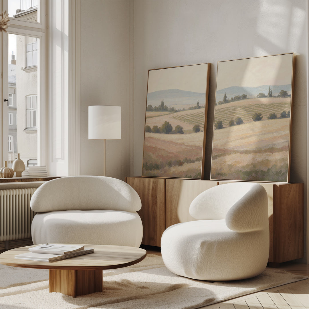 A modern living room with the Harvest Fields - Set of 2 curved white armchairs, a round wooden coffee table, farmhouse decor, and countryside wall art above a sideboard, all illuminated by natural light from large windows.