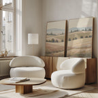 A modern living room with the Harvest Fields - Set of 2 curved white armchairs, a round wooden coffee table, farmhouse decor, and countryside wall art above a sideboard, all illuminated by natural light from large windows.