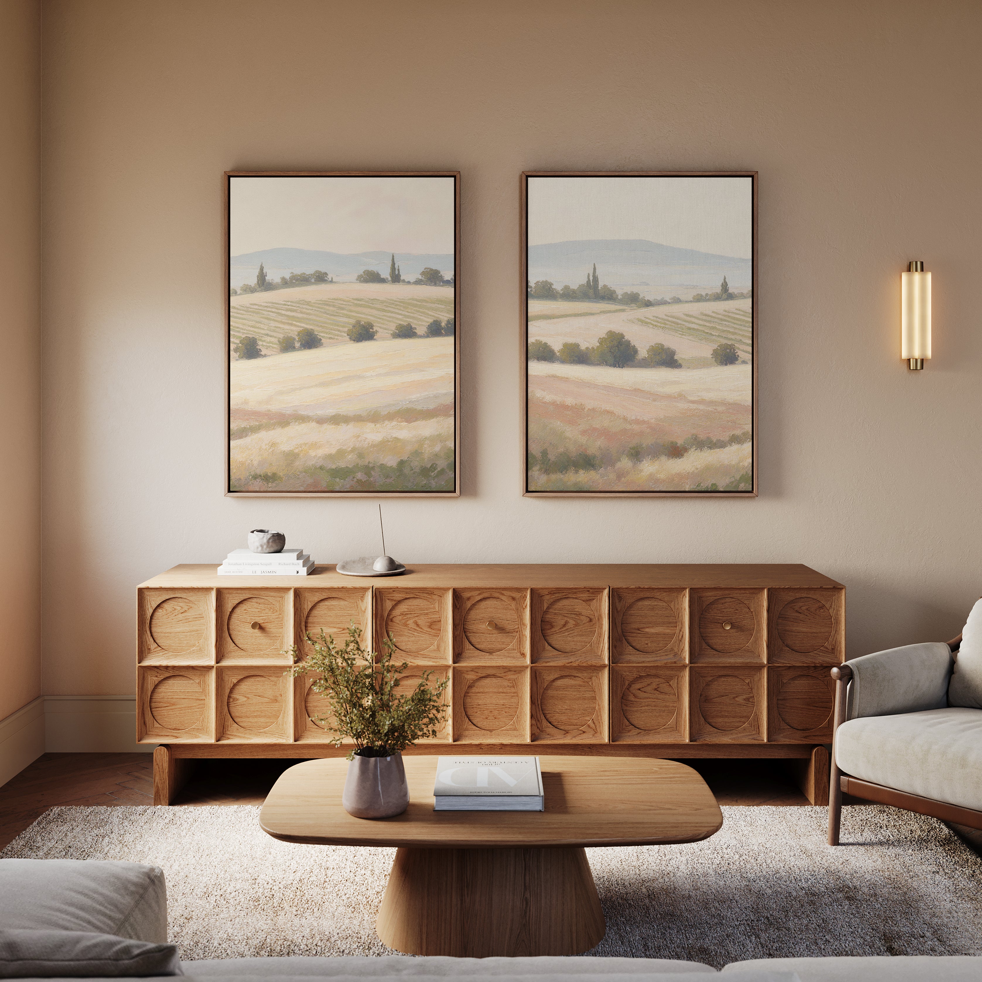 A cozy living room features a carved wooden sideboard, matching coffee table, light armchair, and Harvest Fields - Set of 2 countryside wall art above the sideboard. A small plant on the table completes the serene farmhouse decor.