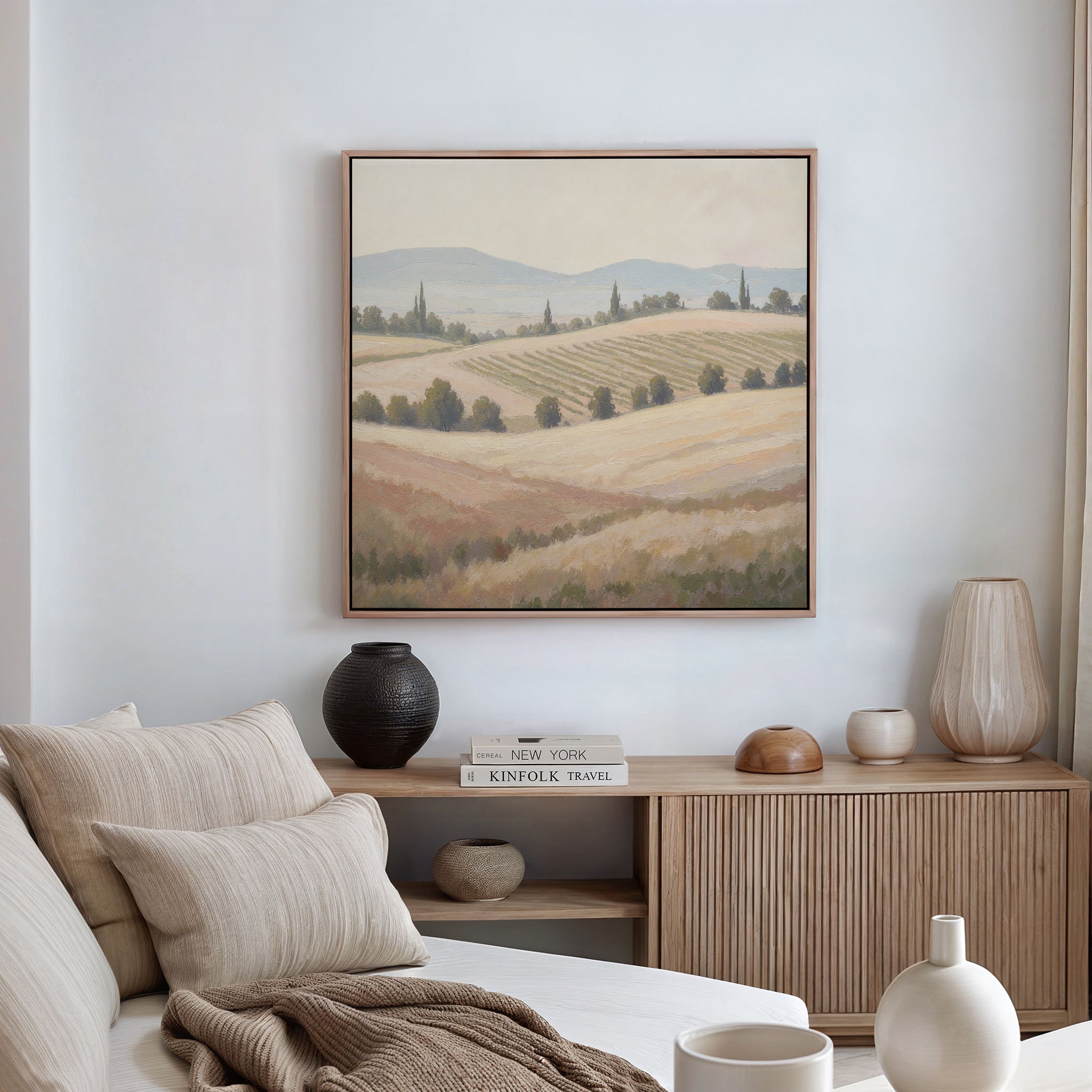 A cozy, neutral-toned living space features a beige sofa with cushions, a wooden sideboard topped with ceramic vases, and the Harvest Fields Canvas Art - Square on the wall. Soft natural light streams in from the right.