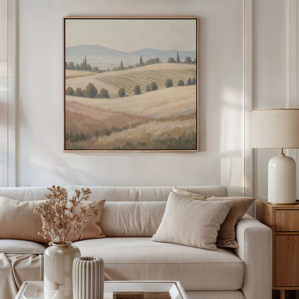 A cozy living room with a beige sofa, neutral cushions, and a lamp on a wooden side table. A vase with dried flowers sits nearby. Above the sofa hangs the "Harvest Fields Canvas Art - Square," enhancing the serene countryside decor.