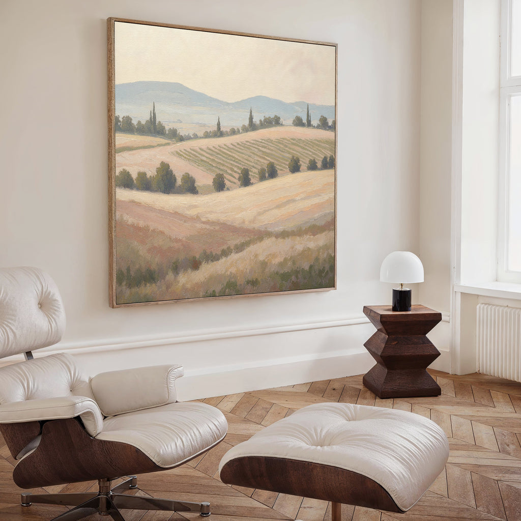 A modern living room with a white leather lounge chair, ottoman, wooden side table, white lamp, and the Harvest Fields Canvas Art - Square above. Light wood floors and natural light complete the stylish countryside decor.