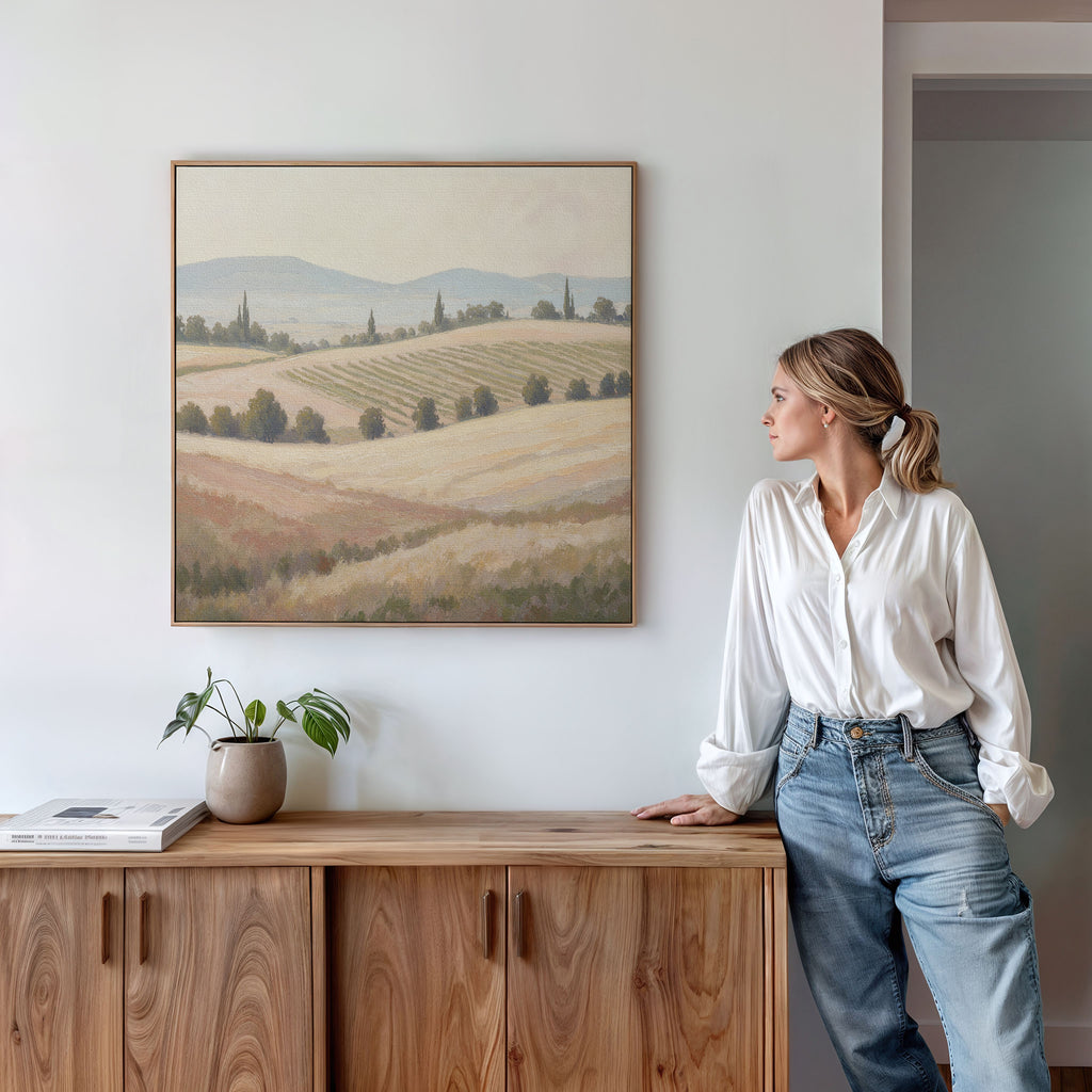 A woman in a white blouse and jeans leans against a wooden sideboard, gazing at the Harvest Fields Canvas Art - Square depicting fields and hills above her. A potted plant and books accentuate the countryside decor on the sideboard.