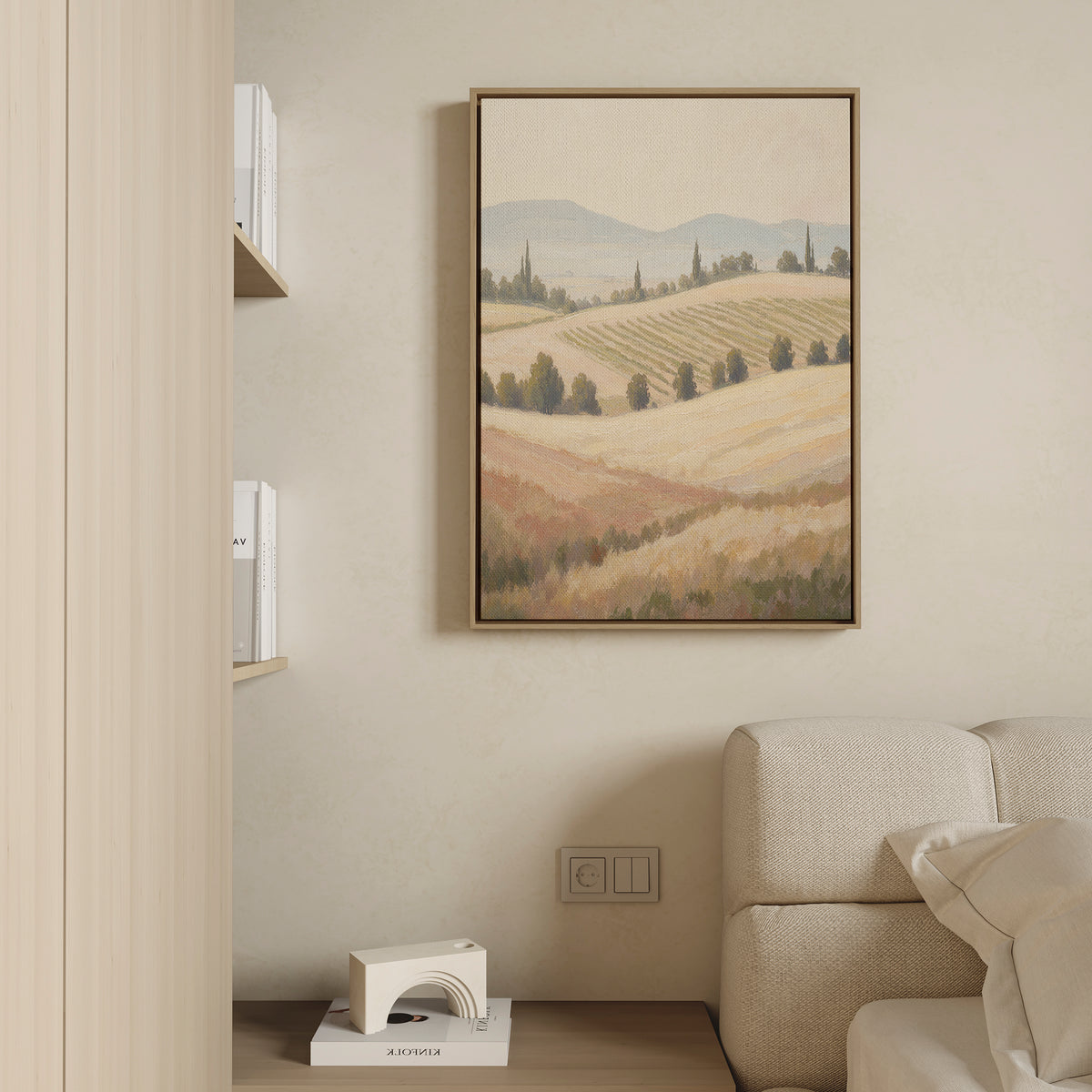 The Harvest Fields Canvas Art - Vertical, featuring rolling hills and trees, hangs above a light sofa on a beige wall. To the left, books and a white geometric decor accentuate the space’s calm, minimalist style.