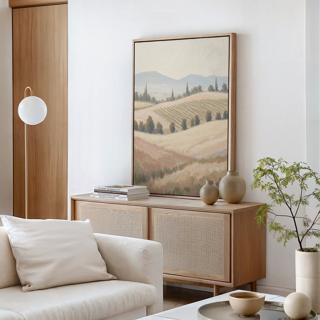 A modern living room with neutral tones features a light wood sideboard, minimalist decor, stacked books, a round white lamp, a potted plant, vases, and the Harvest Fields Canvas Art - Vertical depicting pastoral rolling hills.