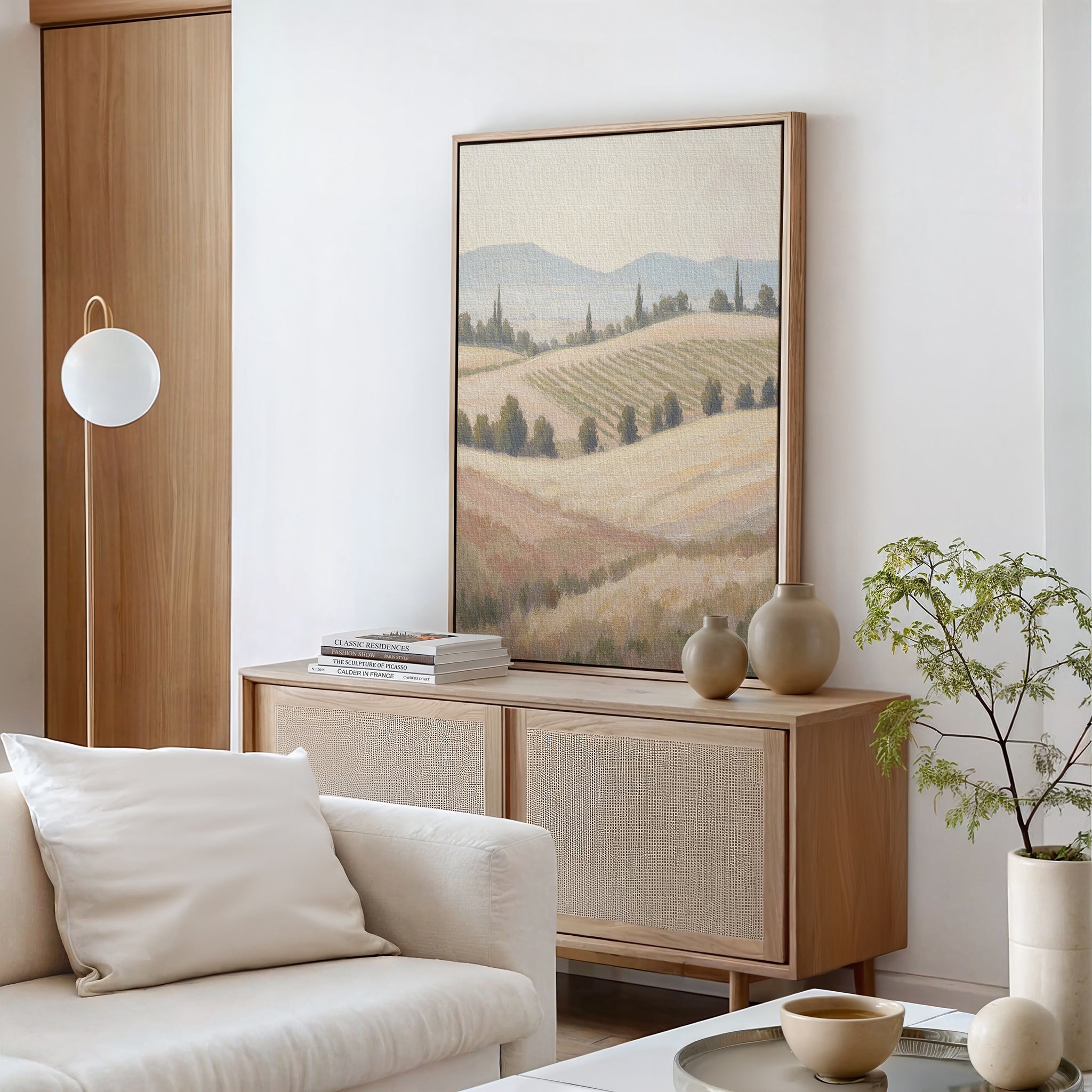 A modern living room with neutral tones features a light wood sideboard, minimalist decor, stacked books, a round white lamp, a potted plant, vases, and the Harvest Fields Canvas Art - Vertical depicting pastoral rolling hills.