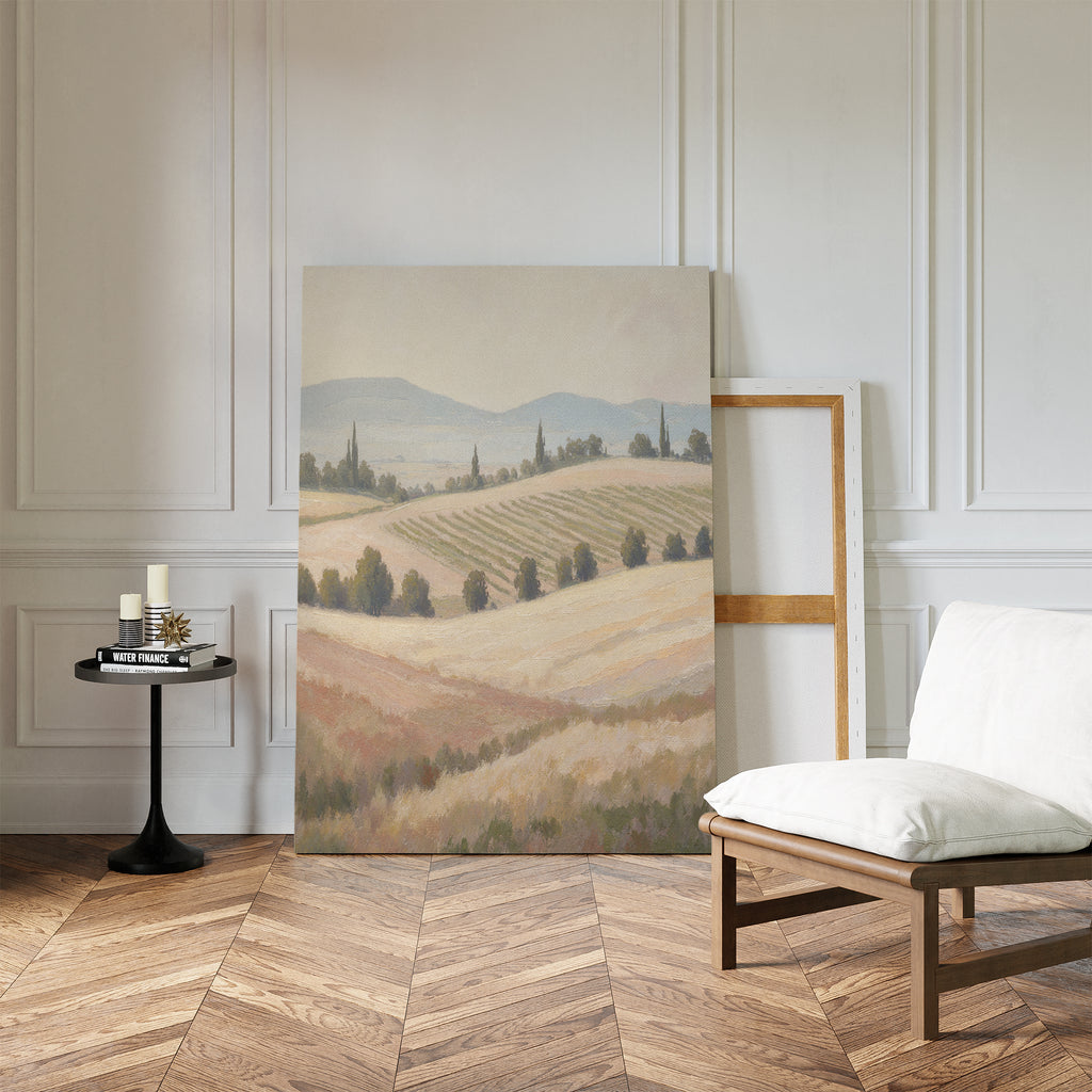 The Harvest Fields Canvas Art - Vertical, featuring a pastoral landscape of rolling hills and fields, leans against a white paneled wall in a bright room with herringbone wood floors, a cushioned chair, and a round table with candles and books.