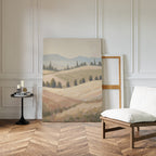 The Harvest Fields Canvas Art - Vertical, featuring a pastoral landscape of rolling hills and fields, leans against a white paneled wall in a bright room with herringbone wood floors, a cushioned chair, and a round table with candles and books.