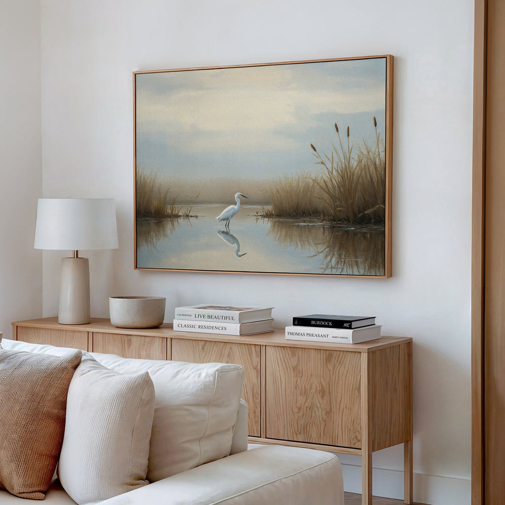 A modern living room features a light wood sideboard, white lamp, and decorative bowls. Above hangs the Heron's Morning Canvas Art - Horizontal, depicting a wetland scene. A beige sofa sits in the foreground.