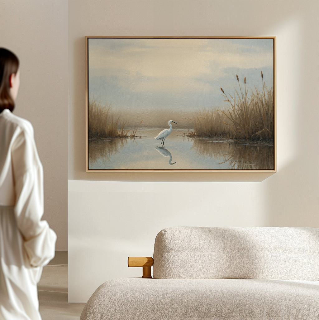 In a minimal, light-toned room, someone in a white robe admires the Heron's Morning Canvas Art - Horizontal: a serene wetland scene featuring a heron standing in calm water among tall grasses.