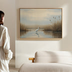 In a minimal, light-toned room, someone in a white robe admires the Heron's Morning Canvas Art - Horizontal: a serene wetland scene featuring a heron standing in calm water among tall grasses.